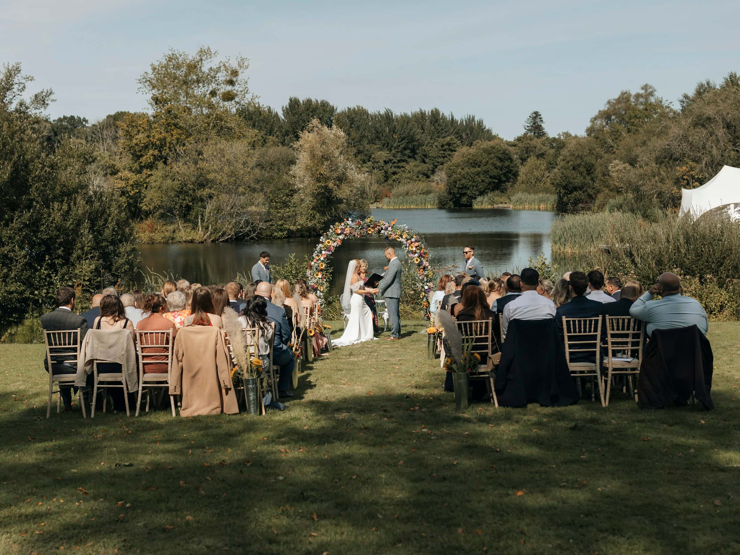 An officiant leads a couple’s ceremony under a floral hoop by a lake