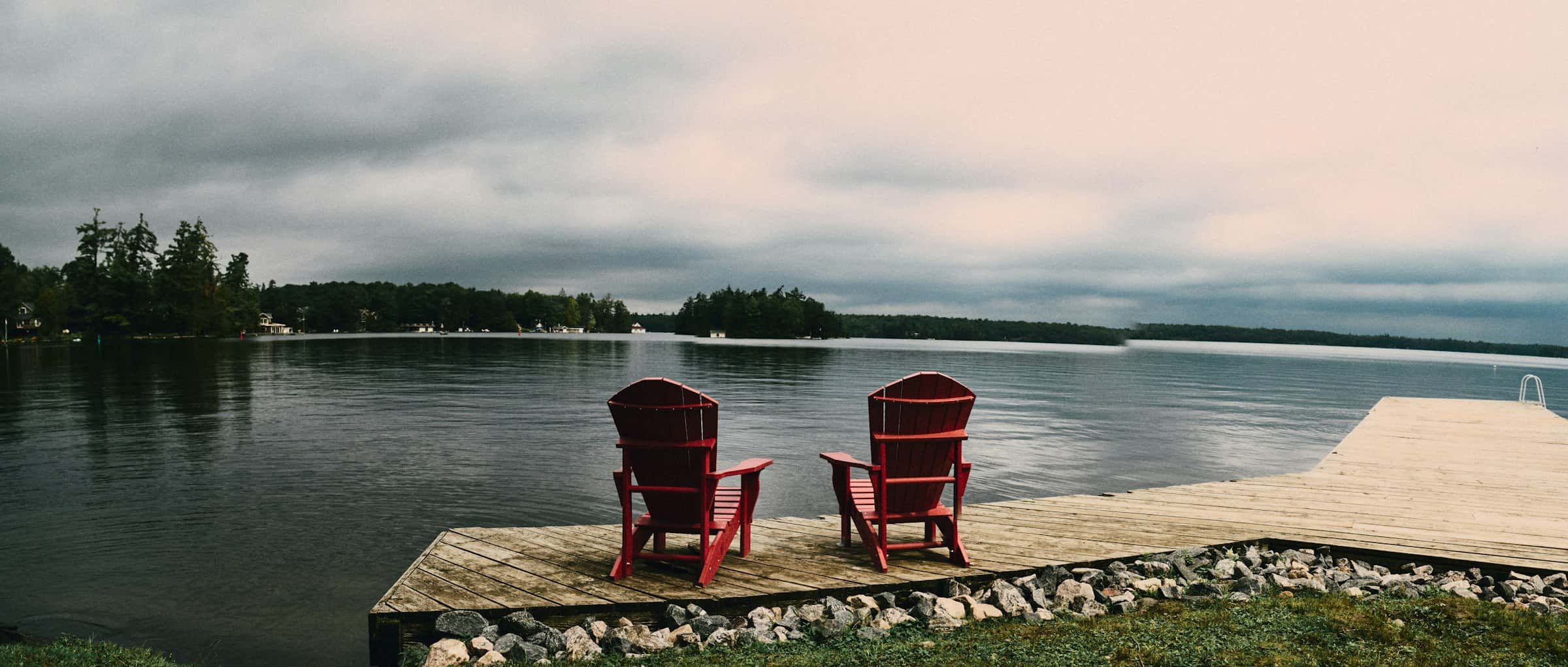 Two red Muskoka chairs on a dock overlooking a lake